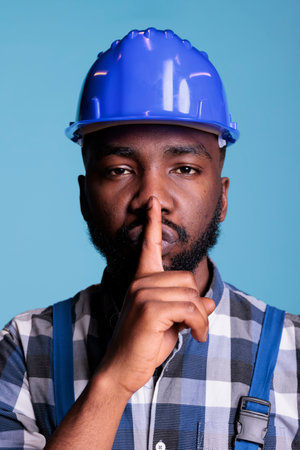 Portrait of male builder showing silence sign in front of camera, putting index finger over mouth to...