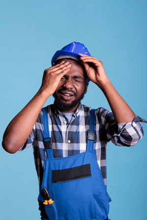Construction worker suffering from a severe headache removing protective helmet. Contractor stressed by overwork touching his forehead with pained expression against blue background.の写真素材
