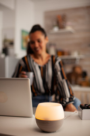 Woman enjoying aromatherapy while working on laptop in kitchen. Aroma health essence, welness home...