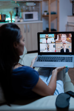 Back view of woman using laptop on videocall sitting on comfortable couch. Remote worker having...