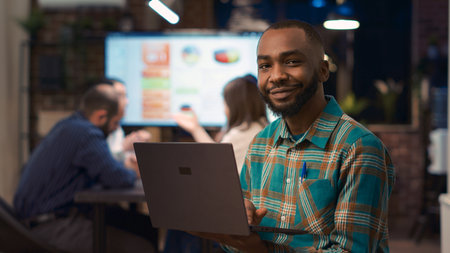 African american employee working on laptop, looking at camera, smiling. Young man holding computer...