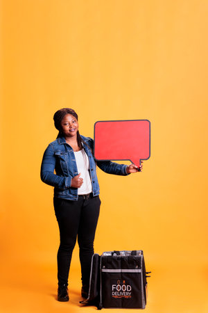 Full body of deliverywoman pointing at red speech bubble advertisting takeaway food service while delivering clients orders. Courier carrying takeaway thermal backpack to deliver mealの写真素材