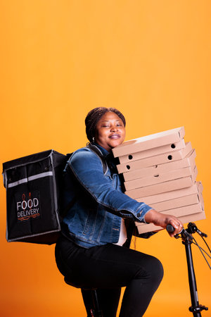 Smiling takeout delivery woman holding carton box stack full with pizza delivering takeaway food order to customer while riding bike. Person standing in studio over yellow backgroundの写真素材