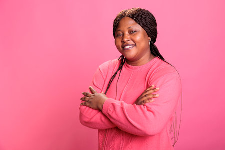 Portrait of excited woman standing in arm crossed while posing in studio over pink background, smiling at camera during shoot. Cheerful model with joyful expression having fun, enjoying free timeの写真素材