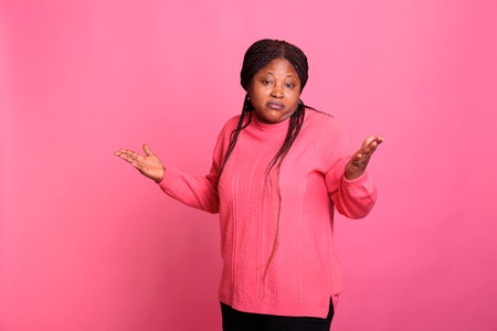 Portrait of doubtful african american woman posing on pink background in studio while making indecisive facial expression. Young adult shrugging shoulders doing i dont know gestureの写真素材