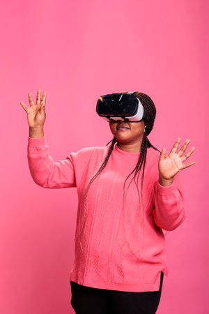Cheerful happy woman wearing virtual reality headset playing video games enjoying 3d entertainment during lesiure time. Happy cheerful person with vr headset while standing over pink backgroundの写真素材