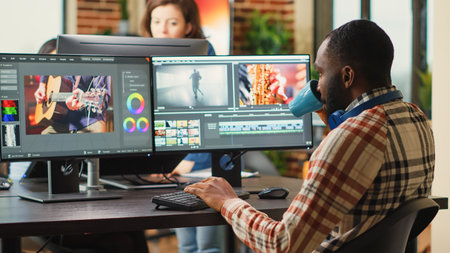 African american employee editing video content on multimedia software, using focus lighting and color grading. Male filmmaker working on movie montage edit on pc multi monitors.の写真素材