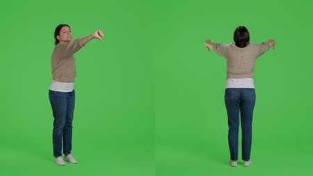 Young adult giving thumbs down negative sign in studio, doing disapproval and bad symbol. Confident woman expressing disagreement with dislike gesture over full body greenscreen.の写真素材