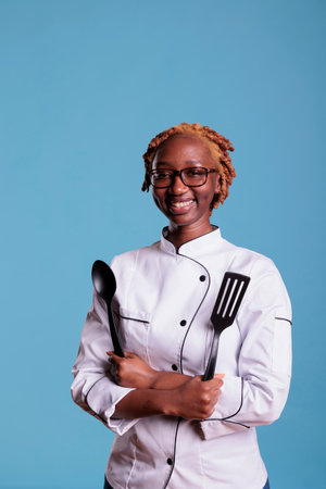 African american female head chef of restaurant holding cooking utensils in studio shot. Female member of kitchen staff with curly hair and uniform jacket smiling against blue background background.の写真素材