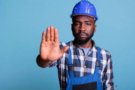 Negative builder doing stop symbol with palm in studio, expressing denial and rejection. Handyman denying and disapproving an idea, raising hand to dismiss and decline suggestion.の写真素材