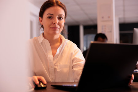 Smiling employee working on laptop in office, looking at camera, marketing specialist using computer...