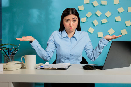 Female employee sitting in modern office making dont know gesture to camera, feeling hesitant and unclear. Insecure professional woman with no idea what should do to finish project.の写真素材