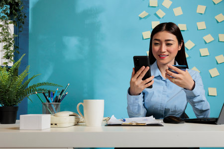 Asian businesswoman holding credit card making financial purchase for business from office. Optimistic female employee making payment transaction at desk using smartphone.の写真素材