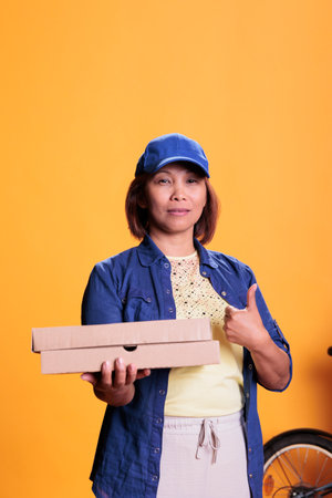 Asian pizzeria delivery employee doing thumbs up gesture while delivering carton flatbox with pizza to customers. Restaurant worker bringing orders with bike. Take out food service and conceptの写真素材