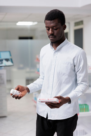 Young african american man deciding between two cardiovascular supplements packages in drugstore, holding heart medication, reading instruction. Buyer thinking, buying prescription treatmentの写真素材