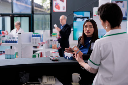Young asian woman asking pharmacy cashier about sunscreen at medical retail store counter desk. Customer shopping in drugstore and consulting with pharmaceutical specialist at checkoutの写真素材