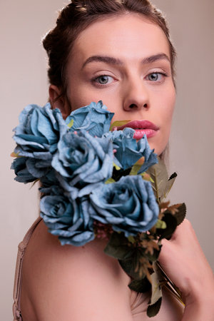 Beautiful woman posing with blue roses bunch on shoulder and looking at camera. Elegant young lady wearing natural make up holding blooming flowers romantic gift in arms close up portraitの写真素材