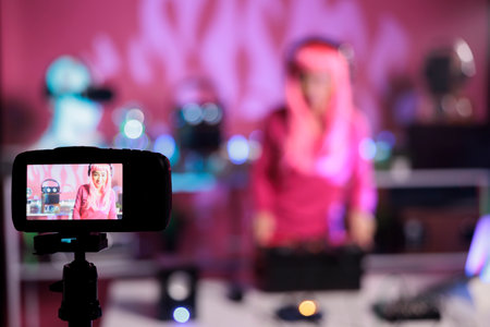 Artist with pink hair standing at table in night club performing techno song using mixer console while filming music session with professional camera. Dj posting performance video on her channelの写真素材