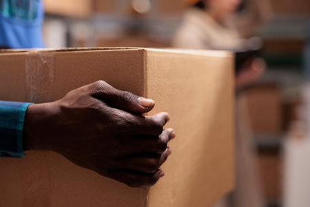 Shipping service warehouse african american man worker holding received parcel in hands. Delivery company employee lifting heavy cardboard box in storehouse with close view on armの写真素材