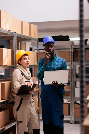 Warehouse workers checking factory goods supply and monitoring dispatching operations on laptop. Caucasian and african american storehouse colleagues standing near cardboard boxes shelfの写真素材