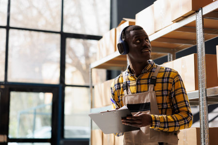 African american storehouse employee in headphones supervising warehouse merchandise. Young industrial warehouse worker listening to music, looking at shelf and taking notesの写真素材