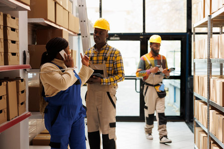 Warehouse manager answering delivery office call and giving instruction to assistant. African american sotrehouse woman operator talking on landline phone and coworker checking order in tablet appの写真素材