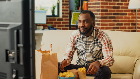 Cheerful person taking bite of cheeseburger on couch, eating burger with fries and bottle of beer....