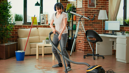 Female adult listening to music on headset and vacuuming floors at home, doing spring cleaning work in apartment. Young wife singing and doing dance moves while she uses vacuum.の写真素材