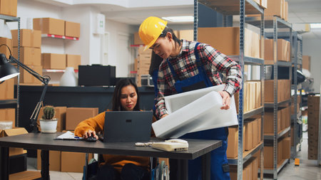 Female wheelchair user checking logistics on laptop in warehouse space, working with employee to send products packages. Person with physical disability doing quality control, small business.の写真素材
