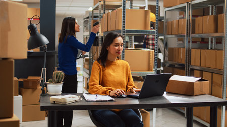 Woman employee analyzing products from racks and shelves, looking at supplies in cardboard packs before shipping order. Young owner working in storage room with business plan.の写真素材