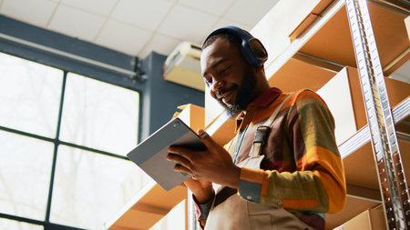 African american man listening to music using tablet, enjoying inventory management in depot with racks and shelves. Male employee having fun dancing on songs, using headphones. Handheld shot.の写真素材