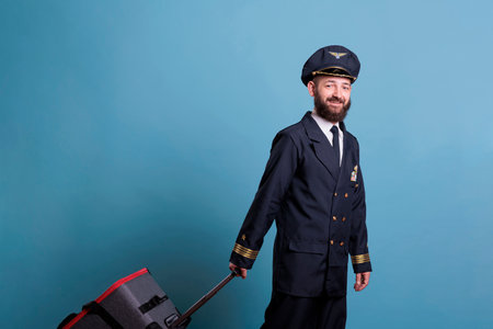 Airplane pilot in professional airline uniform standing with baggage, looking at camera. Ready pilot with luggage waiting for flight in airport, aviator holding luggageの写真素材