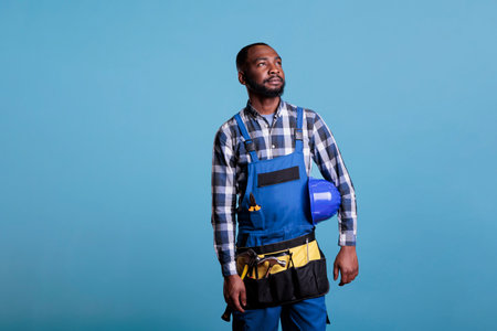 African american construction worker thoughtful looking into distance while holding hard hat under arm. Contractor wearing work clothes and tools against blue background, studio shot.の写真素材