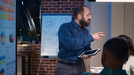 Company financial analyst speaking in business meeting in coworking space, holding digital tablet. Office workers team brainstorming, discussing revenue statistics, looking at graphs on screenの写真素材
