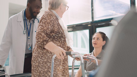 Old person in wheelchair using walking frame at health center, feeling healthy after receiving treatment. Male doctor bringing elderly patient in lobby to meet with little girl. Handheld shot.の写真素材
