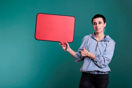 Brunette woman with short hair pointing at empty red speech bubble standing in studio over isolated background. Cheerful female with blank sign for communication on board looking at cameraの写真素材