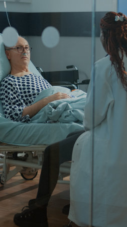 African american doctor talking to sick patient in hospital ward at medical facility. Old man with nasal oxygen tube and oximeter wanting treatment for disease, heavy health problemsの写真素材