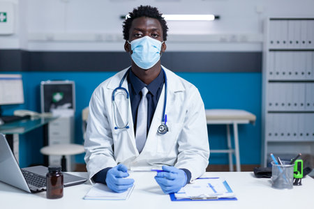 General practitioner in clinic cabinet sitting at desk while wearing facemask because of covid epidemic outbreak. Medic sitting in office while wearing professional uniform and protective gloves.の写真素材