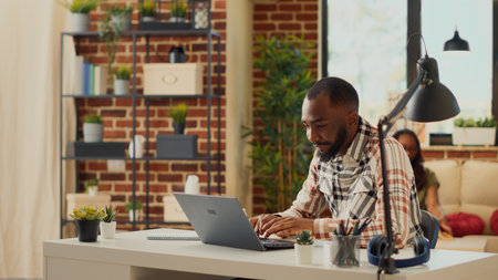 Office manager working from home with laptop, sitting in apartment with partner. Man using computer to plan financial report and send corporate email, work remotely at desk. Tripod shot.の写真素材