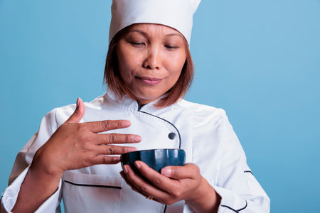 Friendly woman cook smelling healthy dish after cooking culinary meal on blue background. Positive chef wearing restaurant uniform preparing dinner. Gastronomy industry conceptの写真素材