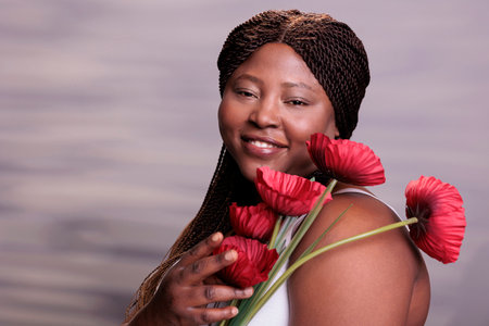 African american woman holding poppy flowers portrait, showing femininity and tenderness concept. Attractive plus size model posing with beautiful summer plants in studio close upの写真素材