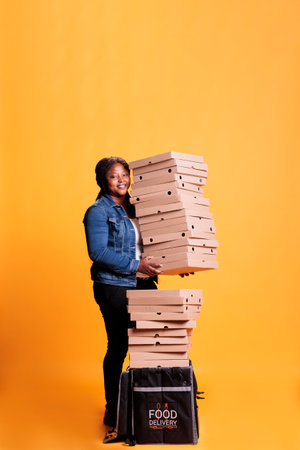 Smiling deliverywoman holding stack full of pizza boxes preparing to deliver to clients during lunch time. Restaurant emloyee with takeout thermal backpack delivering takeaway food orderの写真素材
