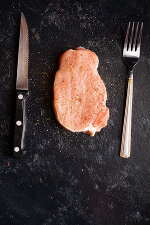 Raw steak meat on black wooden table next to fork and knife. Seasoned piece of meat. Gourmet food and fresh uncooked mealの写真素材