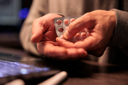 Young programmer taking pills and vitamins from foil, feeling stressed working after hours in app development. Engineer using medicaments and supplements to work late at night. Close up.の写真素材