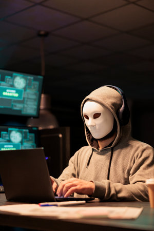Masked scammer hacking computer network at night, working on laptop and multiple monitors to steal IT information. Hacker with hidden identity acting dangerous, server security breach.の写真素材