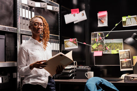 Confident police detective holding crime case file portrait. African american woman law enforcement professional studying investigation documentation, analyzing clues and looking at cameraの写真素材