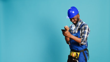 Happy construction worker using smartphone on camera, feeling confident about success. Young smiling carpenter with hardhat browsing online page and texting on mobile phone app.の写真素材
