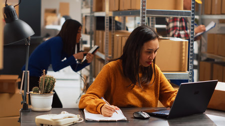 Young woman organizing merchandise in piles, putting retail products in packages for distribution. Female entrepreneur using laptop to check inventory and work on financial planning.の写真素材