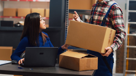 Young man analyzing boxes of supplies in storehouse to help female employee with financial planning and business development. Team of colleagues working on inventory and shipment. Handheld shot.の写真素材