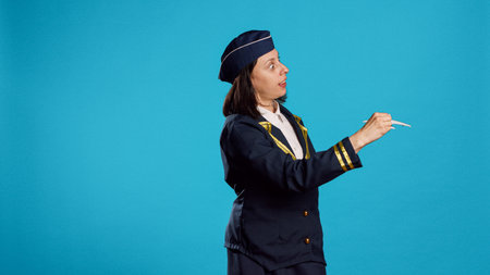 Caucasian woman playing with plane toy, being dressed as stewardess and showing miniature fake airplane in studio. Person working as air hostess holding artificial mini aircraft over blue background.の写真素材